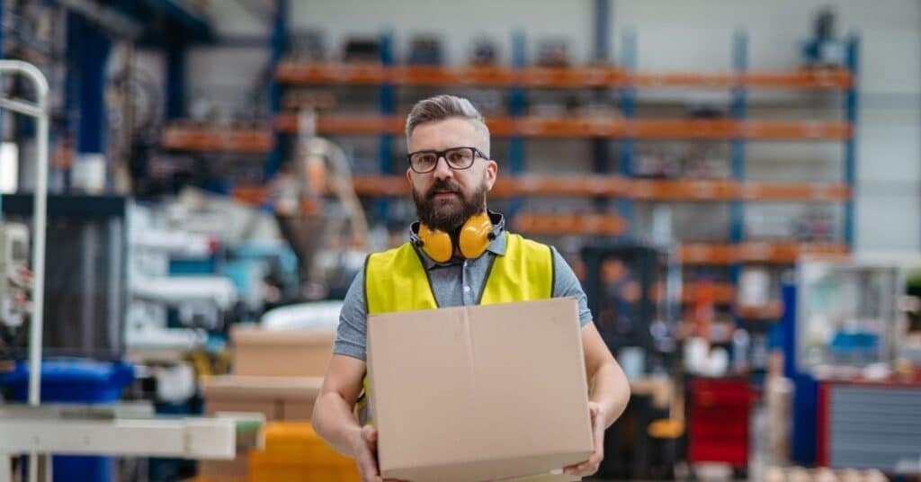 a middle aged Caucasian man is carrying a box in a warehouse and wearing orange ear protection around his neck